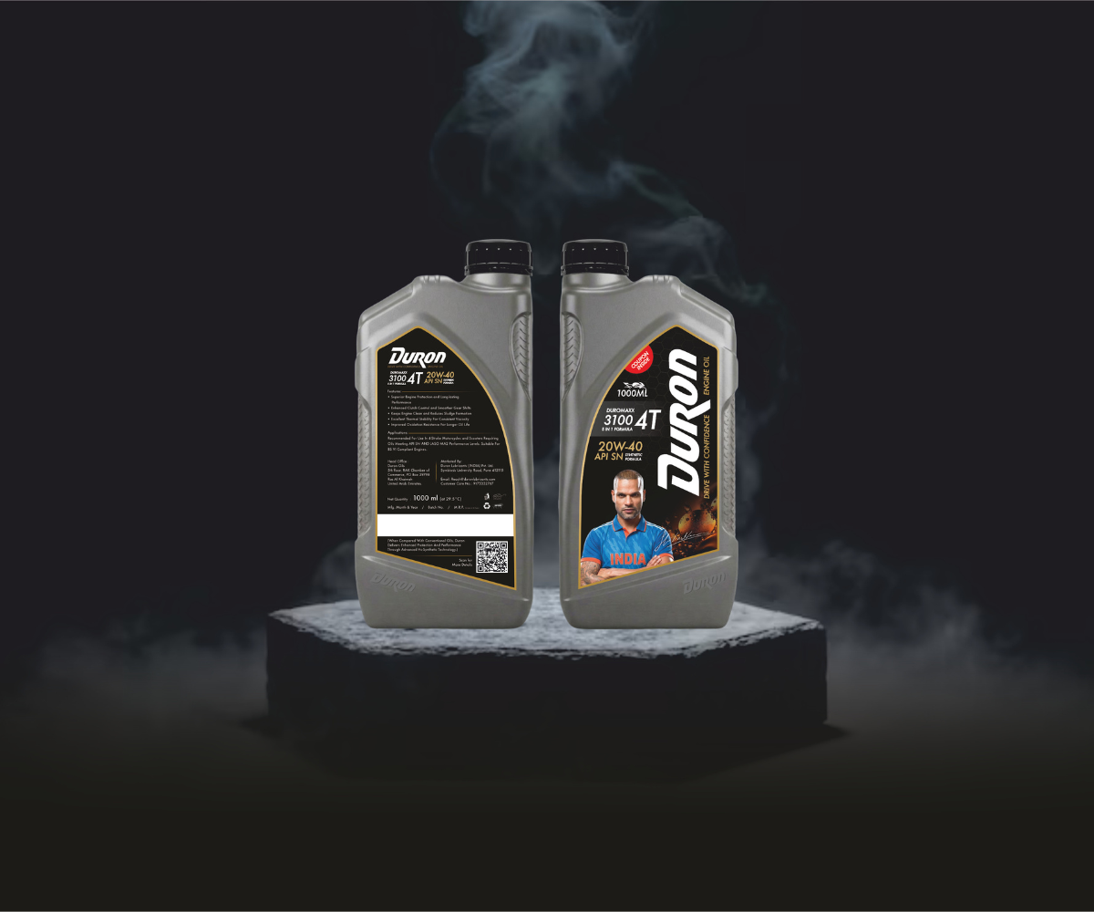 Duron 3100 4T 20W40 synthetic motorcycle engine oil, 900ml grey bottles with black caps, front and back labels visible, cricketer image (Shikhar Dhawan in blue jersey), QR code, black-gold design, displayed on a dark smoky background.
